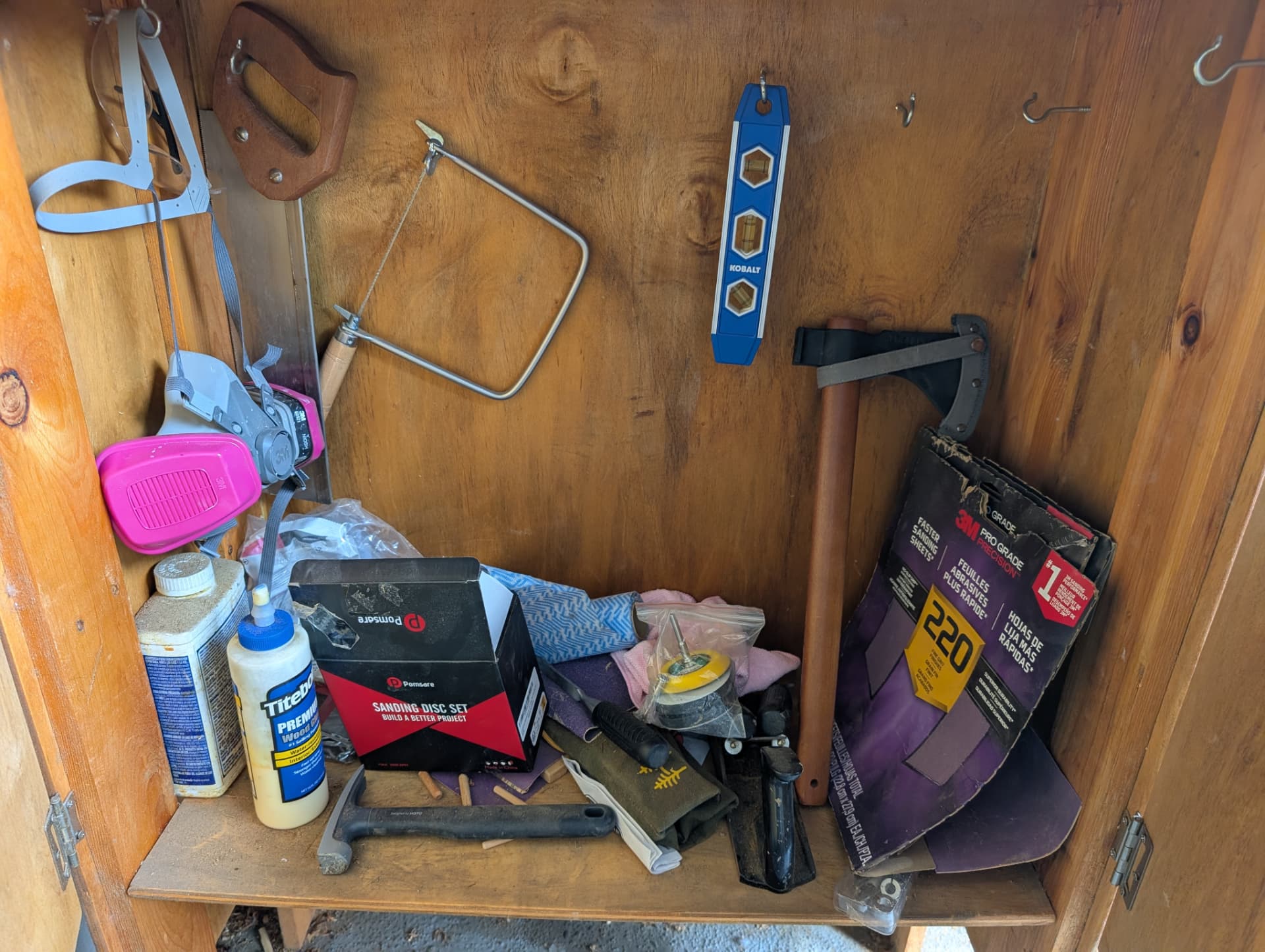 Homemade wooden tool cabinet holding hand saws, a coping saw, level, clamps, wood glue, respirator, and sanding supplies